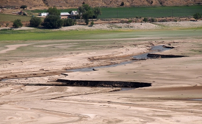 [Some water sits in the middle of a series of ridges in the dirt that is normally the bottom of the reservoir. Grass and some buildings are seen in the distance.]